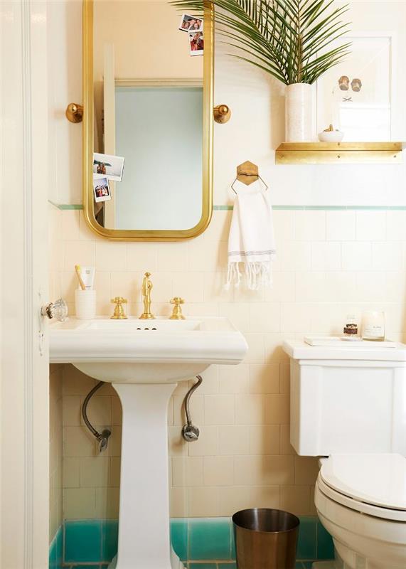 įrėmintas veidrodis, aukso spalvos čiaupai, baltas baseinas, maža lentyna, vaza su egzotišku lapu, wc apdaila