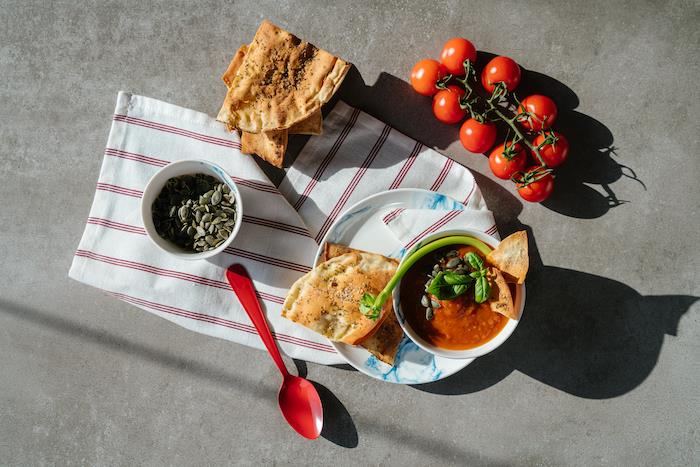 pomidorų sriuba, keraminis dubuo, pilnas sriubos, pomidorų ir sėklų, išdėstytas ant pilko stalviršio, apačioje raudonas ir baltas audinys