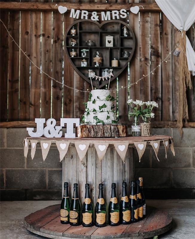 deko medžio ritė, maražo pyrago stalo idėja, šalies prašmatnumas, baltas pyragas ant medinio rąsto, vedusios didžiosios raidės, gėlės stikliniuose buteliuose, šampano buteliai