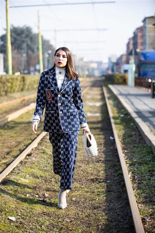 ženska, ki hodi po vlakovnih tirih, trendi v slogu 2019, nosi modro obleko s hlačami in blazerjem, bele pete in torbo