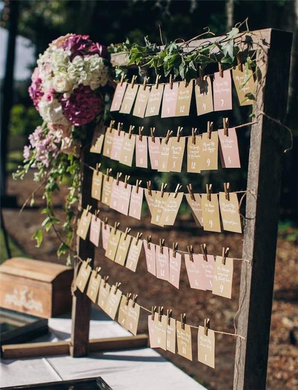vestuvių stalo planas dideliame kaimiškame mediniame rėme su svečių vardų etiketėmis ir spalvotu popieriniu stalu, žalia girlianda ir spalvinga gėlių puokšte