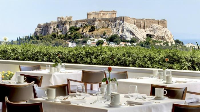 žalia siena kaip turėklas restorane Atėnuose, šviesiai rudos kėdės, gražus vaizdas į Akropolį, be debesų mėlynas dangus