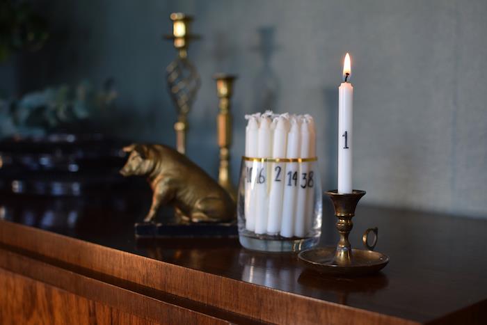 originali ir paprasta suaugusiųjų advento kalendoriaus idėja baltomis sunumeruotomis žvakėmis su juodais skaičiais ant senovinės komodos