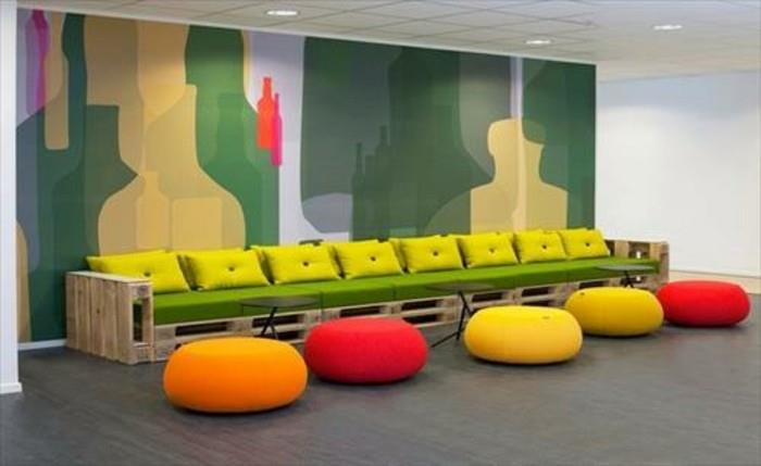 ilgas suoliukas arba padėklų sofa, pagaminta iš šviesių medinių padėklų, padengta žaliu putų čiužiniu ir dekoruota ryškiomis geltonomis pagalvėlėmis