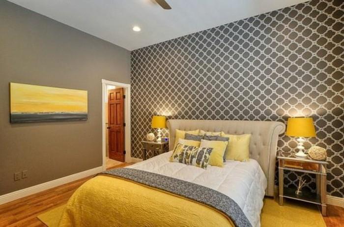 tapyba geltonos ir pilkos spalvos, pilkos spalvos miegamojo viduje, su raštuotais tapetais, šviesiai smėlio spalvos lova, balta ir pilka bei geltona patalynė