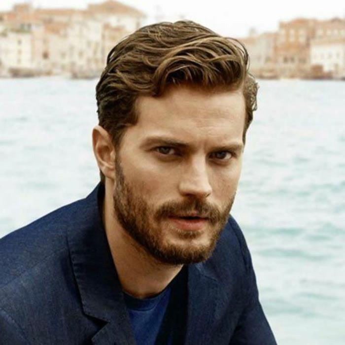 jamie dornan z valovitimi, kratkimi lasmi in česami, počešanimi na eno stran, brado in brki, temnim denim blazerjem in modro majico