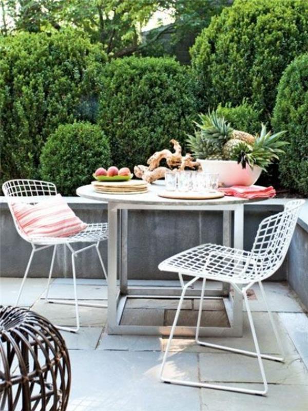 meyve ve süslemeli yuvarlak metal masa ve biri pembe çizgili yastıklı iki beyaz sandalye, dış mekan veranda fikirleri, arka planda yeşillik