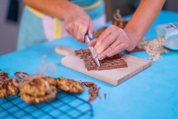moteris, laikanti peilį, pjaustanti šokoladą į gabalus, kramtomus šokoladinius sausainius, mėlyną stalą, medinę pjaustymo lentą