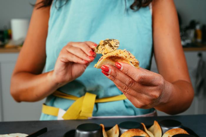 moteris su raudonu nagų laku, užpildo tortilijos įvyniojimus, taco mėsos receptą, žalvarinį šaukštą, mėlyną prijuostę