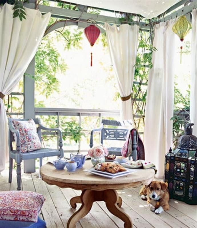 kapalı veranda fikirleri, çay seti ve hamur işleri ile yuvarlak ahşap masa, iki soluk mavi süslü sandalye, kapalı veranda fikirleri, beyaz perdeler ve bir köpek