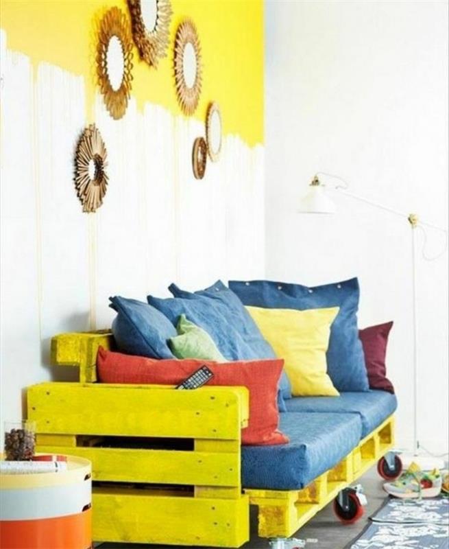 ryški geltona sofa, pagaminta iš dažytų padėklų, pritvirtinta kartu ir pridedama ratų, kaip pagaminti padėklų baldus, dekoruotus pagalvėmis mėlynos, raudonos ir geltonos spalvos