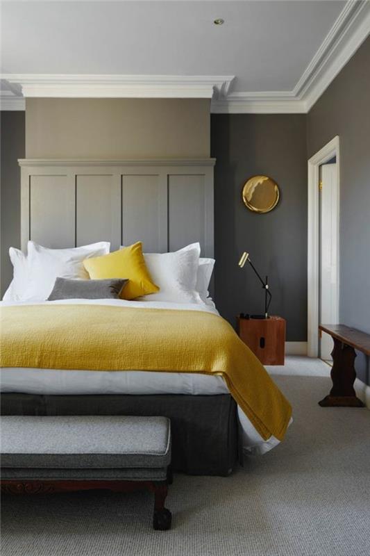 suoliukas tamsiai rudame, kreminės pilkos spalvos miegamajame su dailylentėmis, šviesiai pilku kilimu, lova su balta ir pilka spalva, geltonais užvalkalais ir pagalvėmis